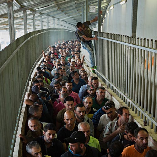 Palestinian workers line up in order to pass the Israeli checkpoint in Bethlehem, on the West Bank; some climb up the fence, forming a parallel line in midair. Photo by Lalo de Almeida/Folhapress
